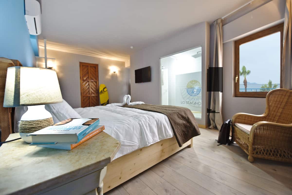Una habitación de hotel con una cama, una mesita de noche con un libro, una lámpara, una silla de mimbre y una ventana con vista al mar.