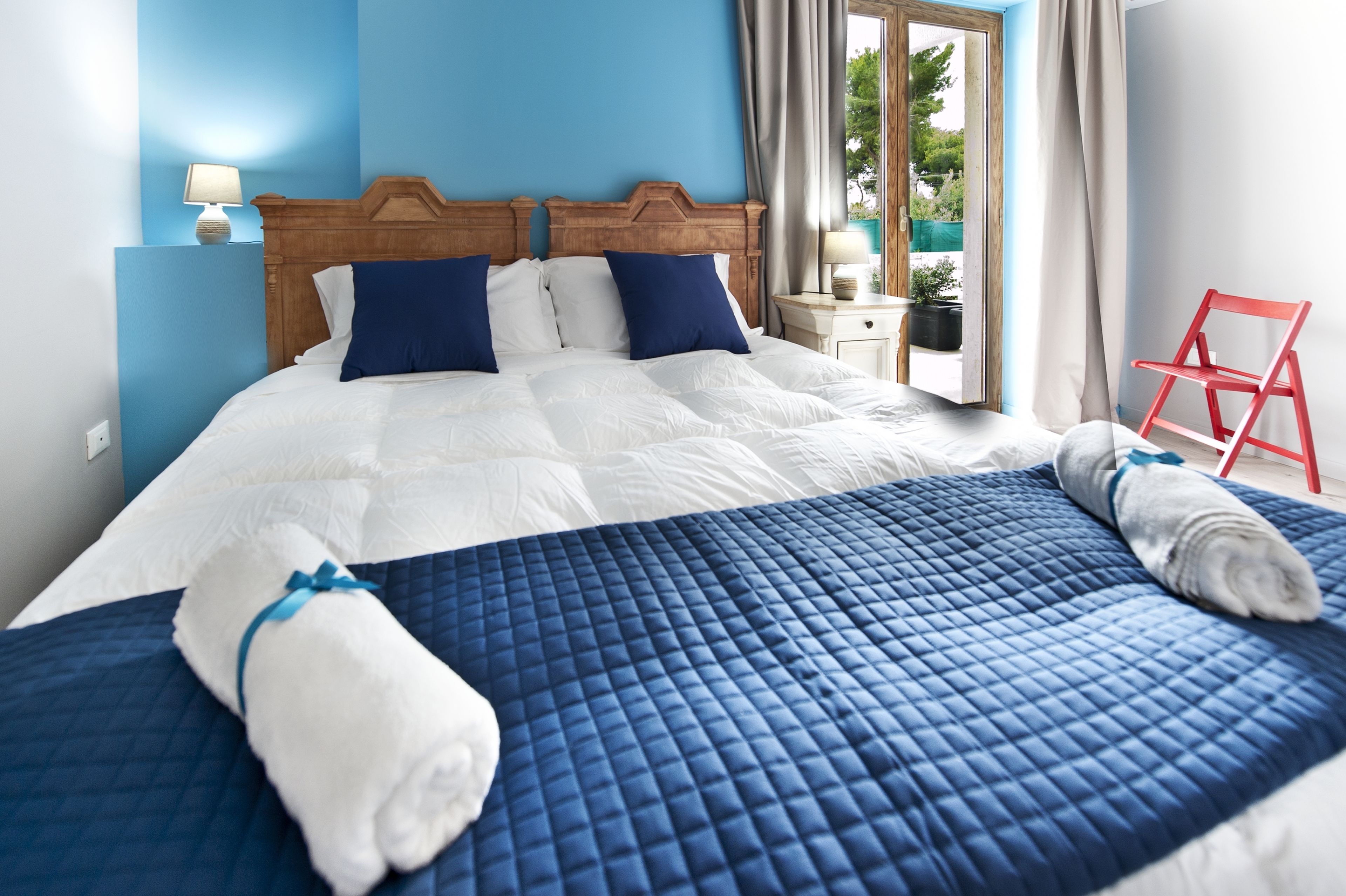 A neatly made bed with a blue quilt and white linens, a wooden headboard, and a red chair in a room with blue walls and a window.