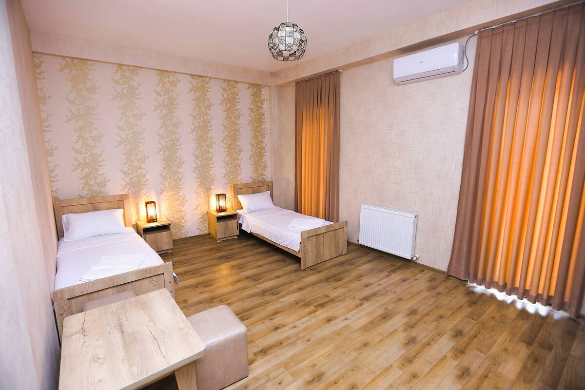 Una habitación con dos camas individuales, un banco de madera, una mesita de noche con lámpara y un aire acondicionado instalado en la pared.