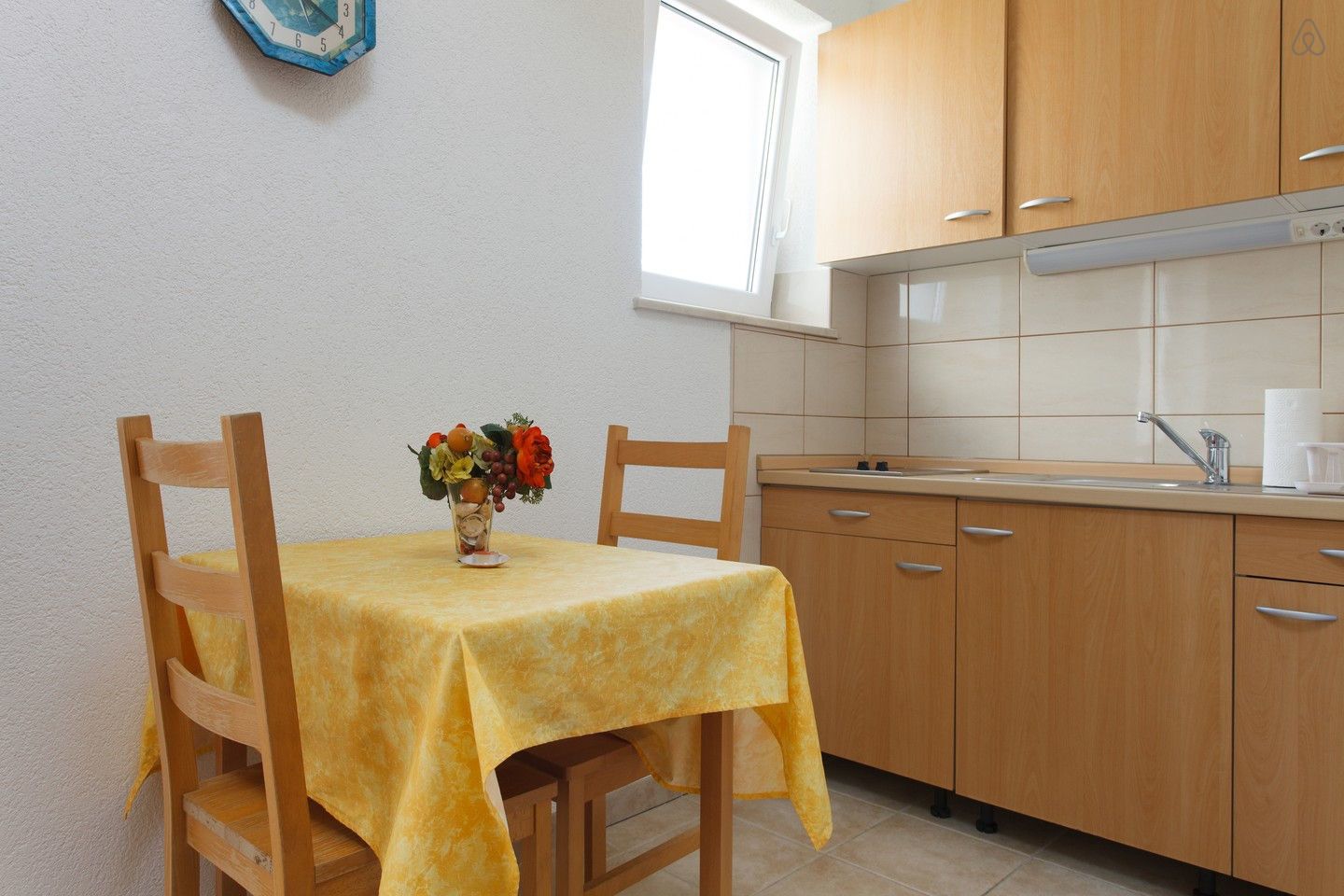A small kitchen with wooden cabinets, a window, and a table with a yellow tablecloth and a fruit vase.