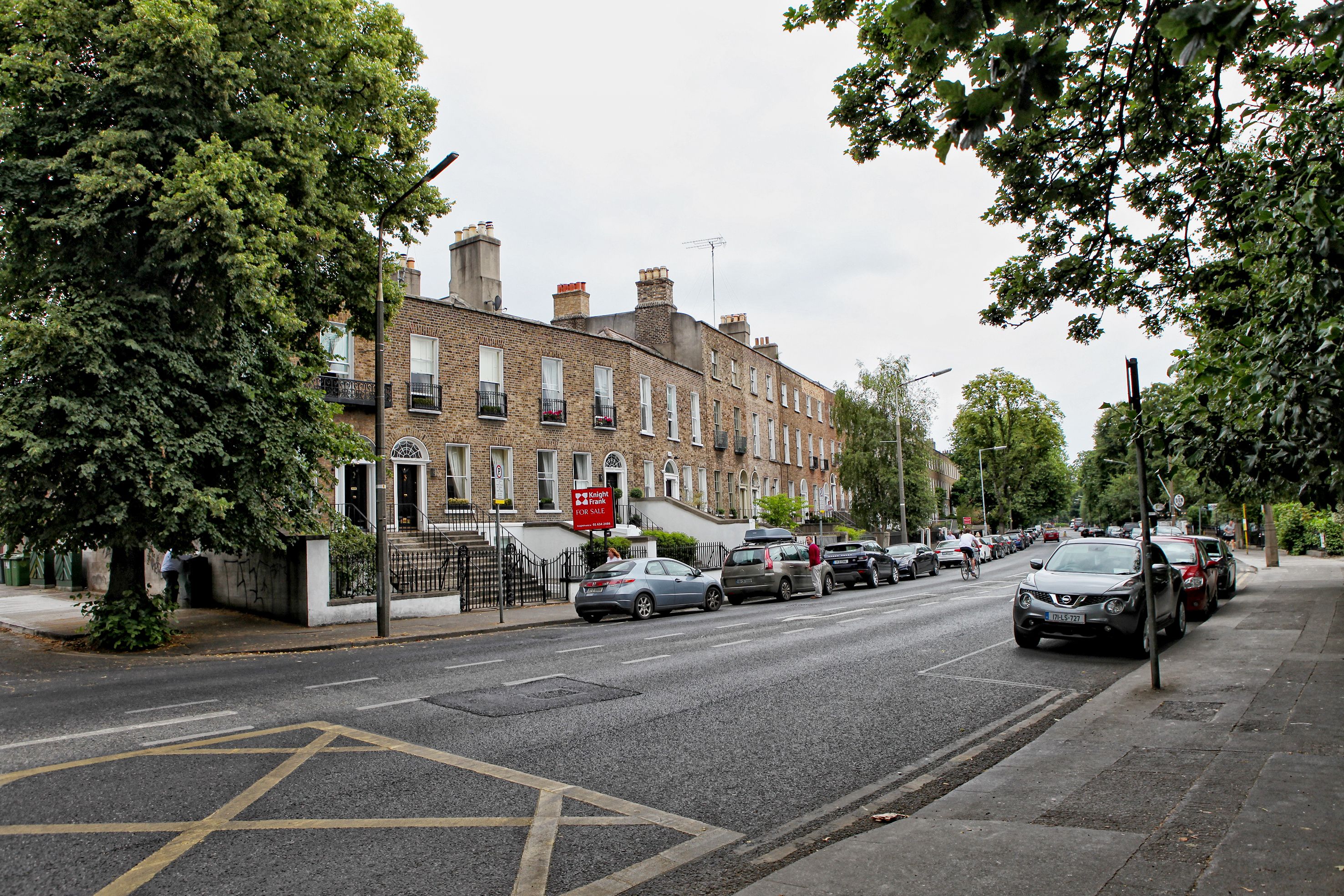 A street lined with brick townhouses, parked cars, and a 'For Sale' sign.
