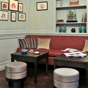 A living room with a red sofa, two dark tables, and a coffee table. There are framed botanical prints on the wall and decorative vases on a shelf.