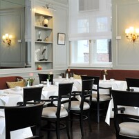 Een restaurant met meerdere tafels en stoelen, witte tafellaken en uitzicht buiten door grote ramen.