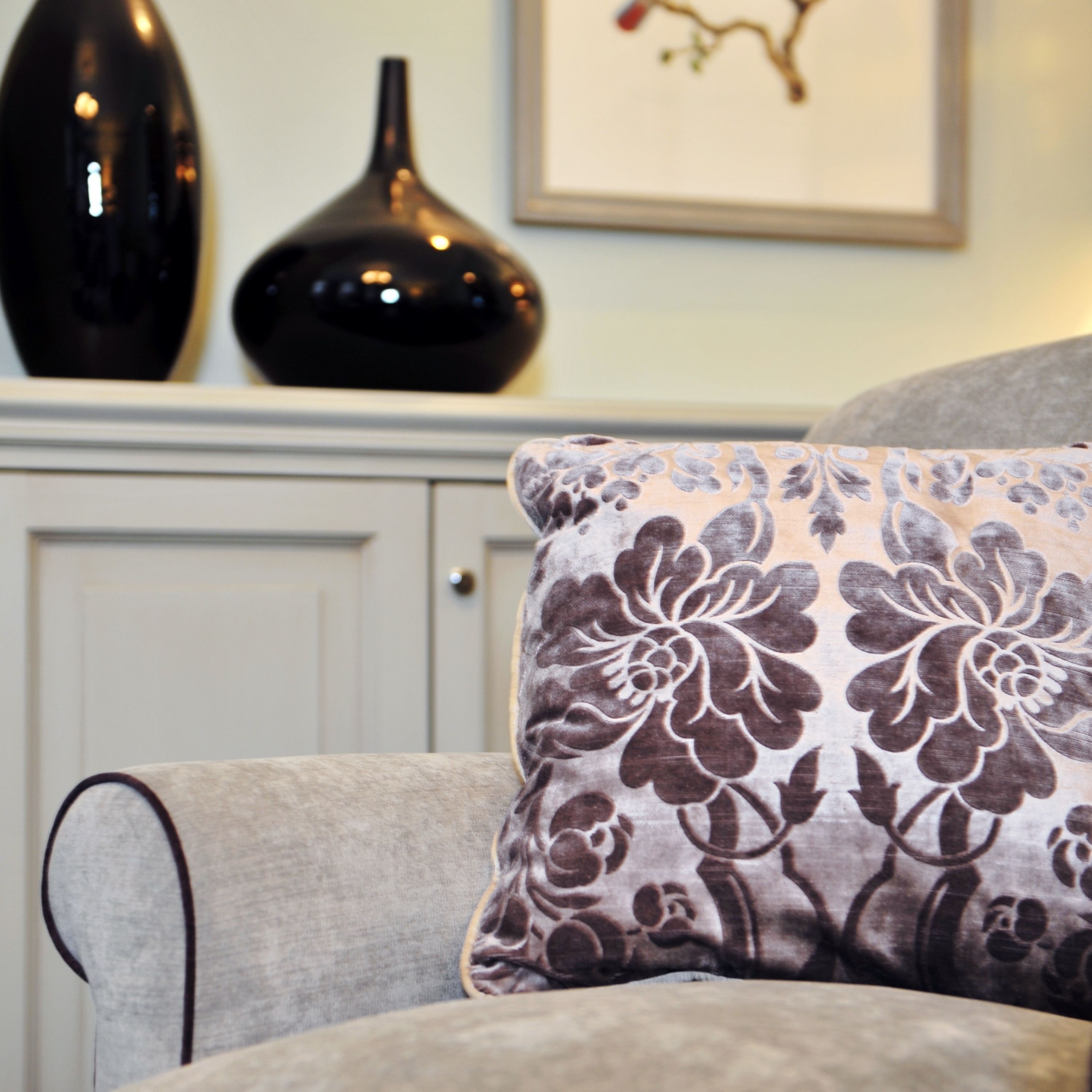 A sofa with a floral-patterned cushion, a black vase, and a framed picture on a wall.