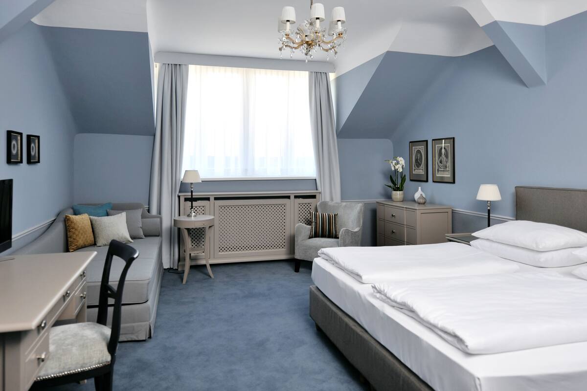 A hotel room with a large bed, a desk, a chair, a small table, a radiator, and a window with curtains.