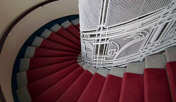 Escadaria em espiral com degraus revestidos de carpete vermelho e grades de metal branco.