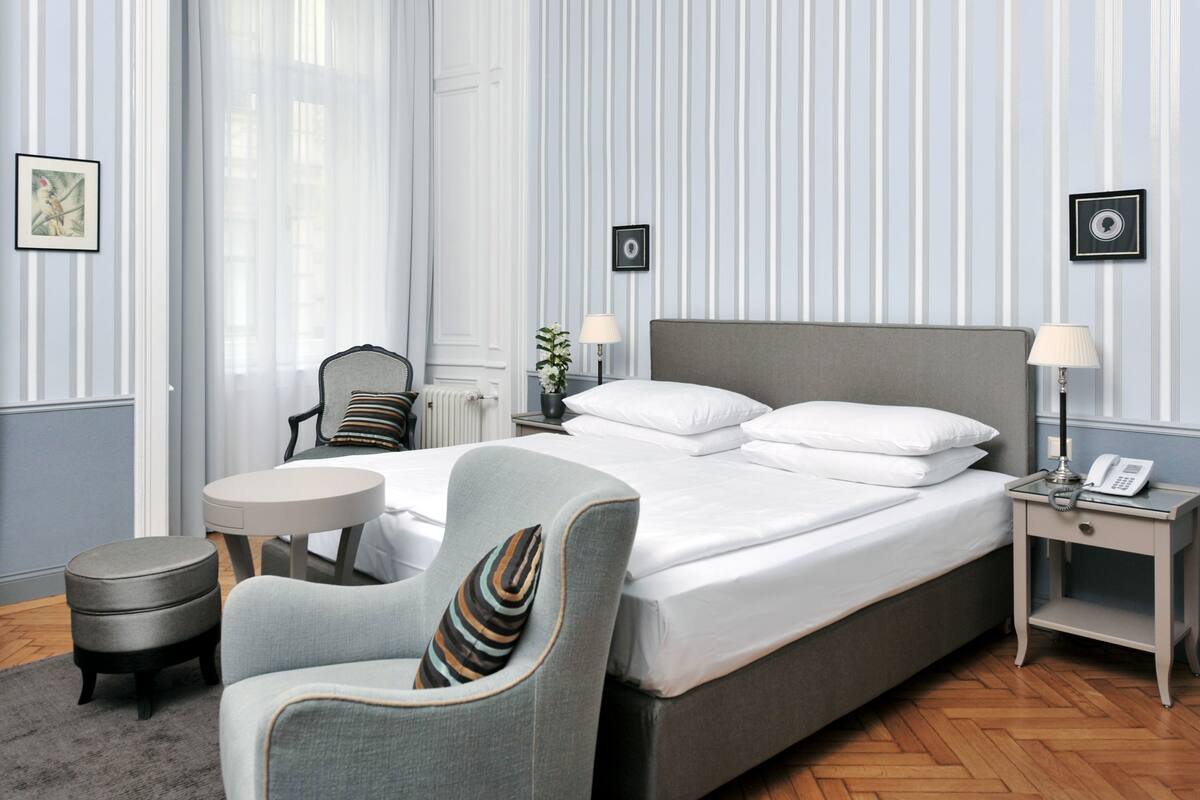 A hotel room with a large bed, a chair, a small table, and a nightstand with a lamp and phone.