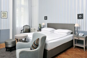 Quarto de hotel com cama grande, uma cadeira, mesa pequena e mesa de cabeceira com abajur e telefone.