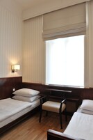 Quarto de hotel com cabeceira de madeira, mesa de cabeceira com abajur, um banco e uma janela grande com cortinas.