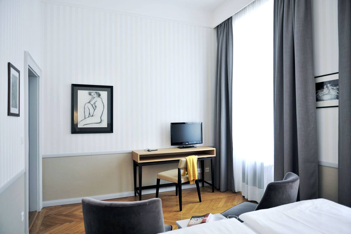 A hotel room with a bed, a desk with a television, a chair, and a window with curtains.