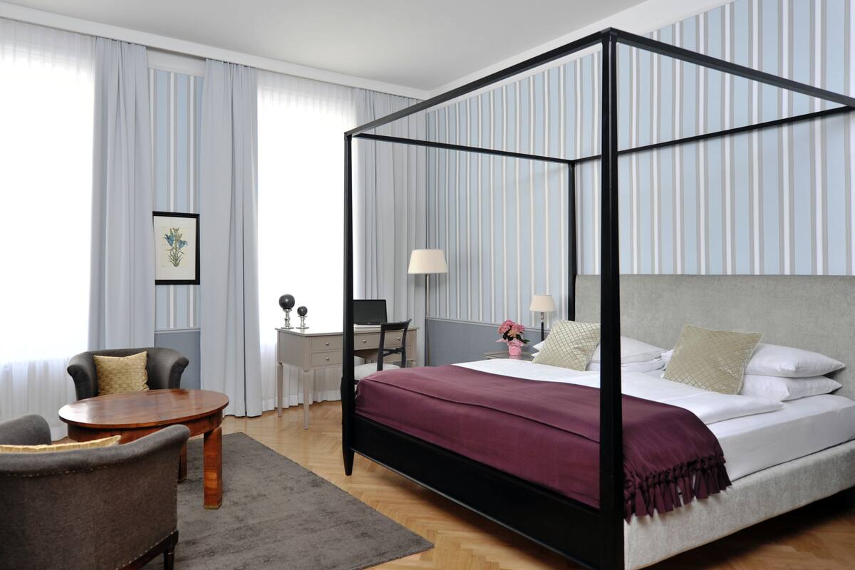 A four-poster bed with a headboard, a desk with a chair, a round wooden table, and a grey rug in a hotel room.
