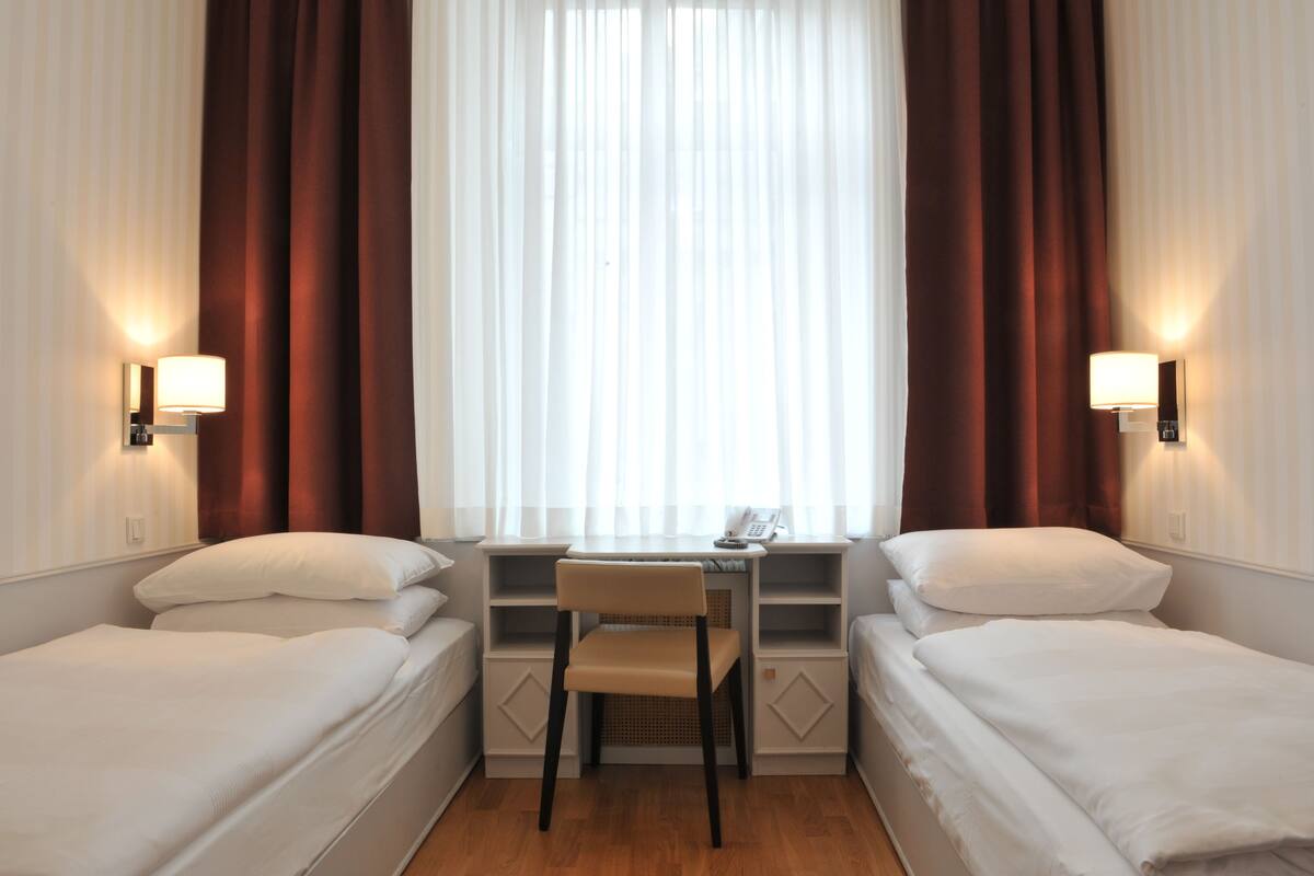 A hotel room with two single beds, a small desk with a chair, and a window with curtains.
