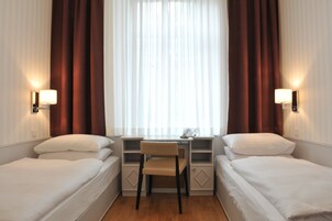 Quarto de hotel com duas camas de solteiro, uma secretária pequena com cadeira e uma janela com cortinas.