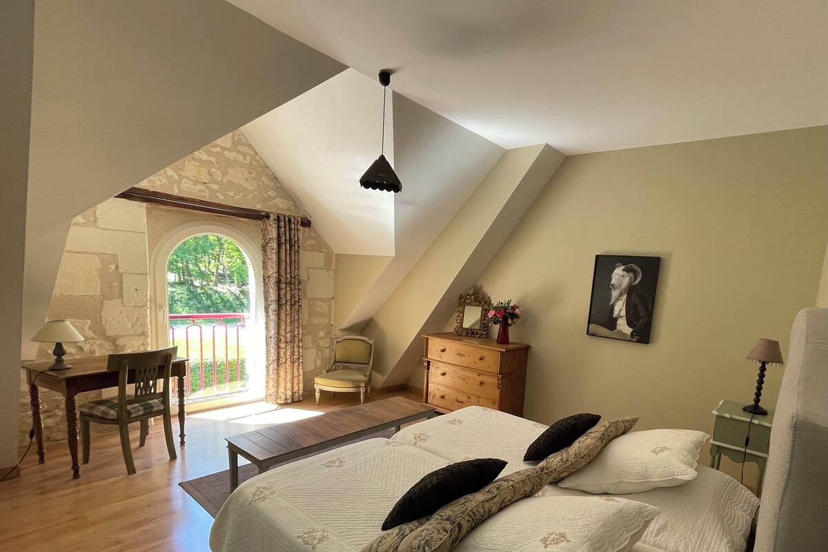 Ein Schlafzimmer mit schrägen Wänden, einem Bett mit floralem Bettzeug, einem Schreibtisch mit Stuhl und Blick auf Grünfläche.