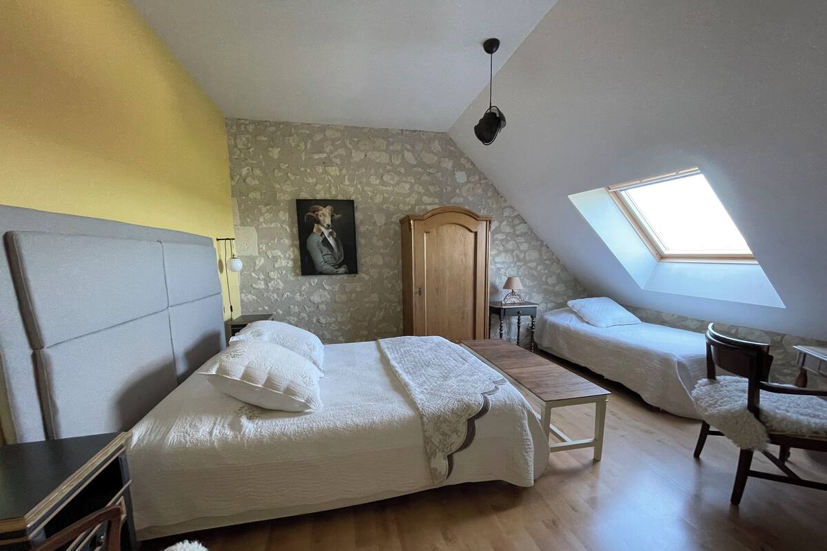 Ein Schlafzimmer mit einem großen Bett, einem kleineren Bett, einem Holzschrank, einem Schreibtisch und einem Stuhl. Es gibt ein Dachfenster und eine Steinwand.