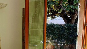 A bedroom with a large bed, a wooden door leading to a garden, and a view of a tree and hedge outside. - Louminai (Donzere)