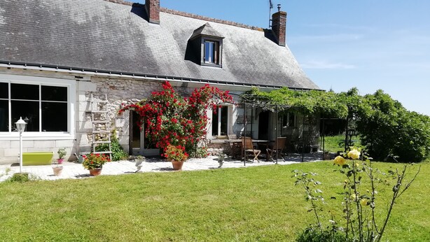 Une maison en pierre avec un toit en pente, une petite fenêtre et un jardin agrémenté de plantes en pot et d’une pergola.