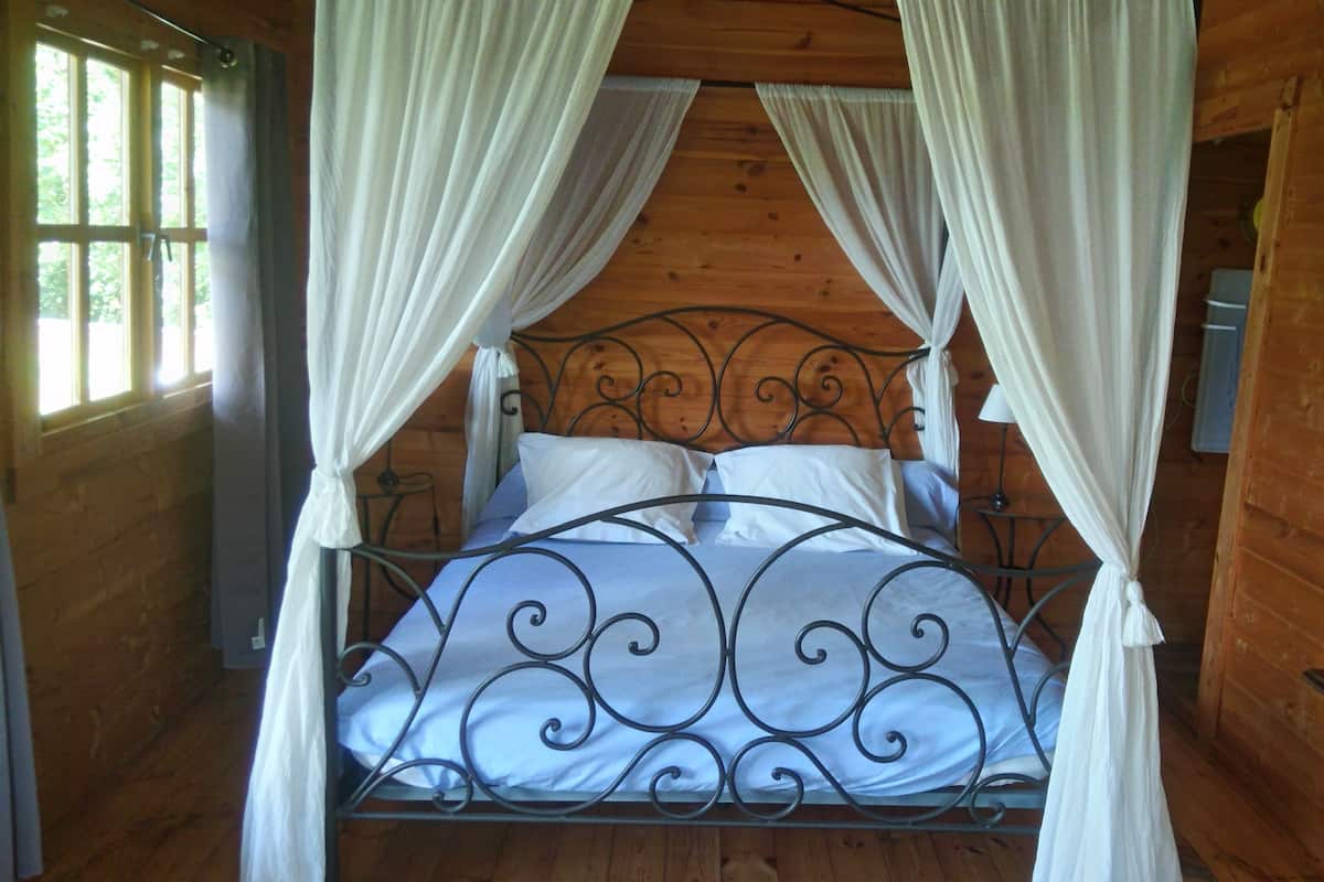 Ein Schlafzimmer einer Holzhütte mit einem Metallbettgestell, weißen Vorhängen und einem Bild an der Wand.