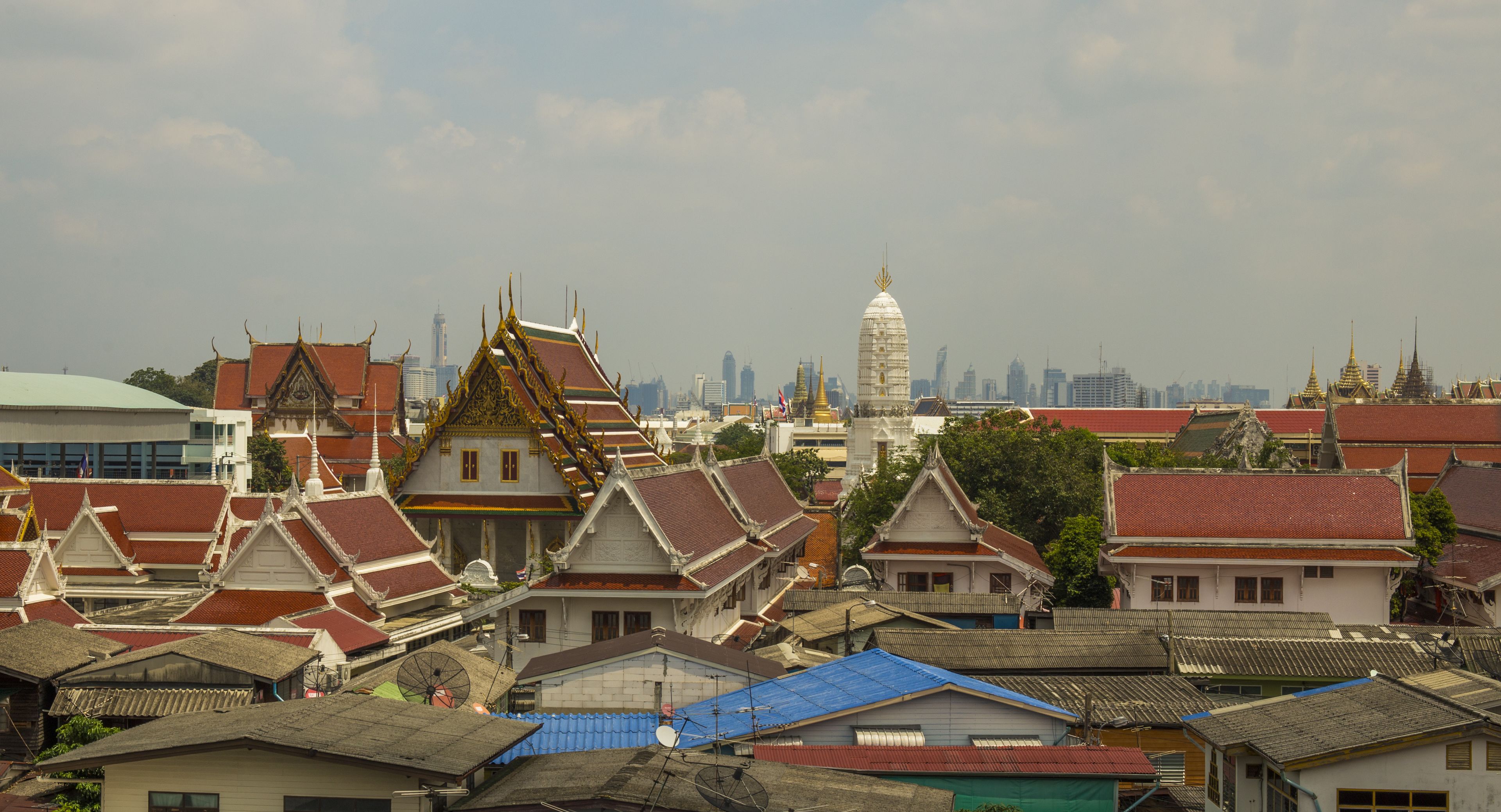 A traditional Thai temple complex with ornate architecture and a city skyline in the background.