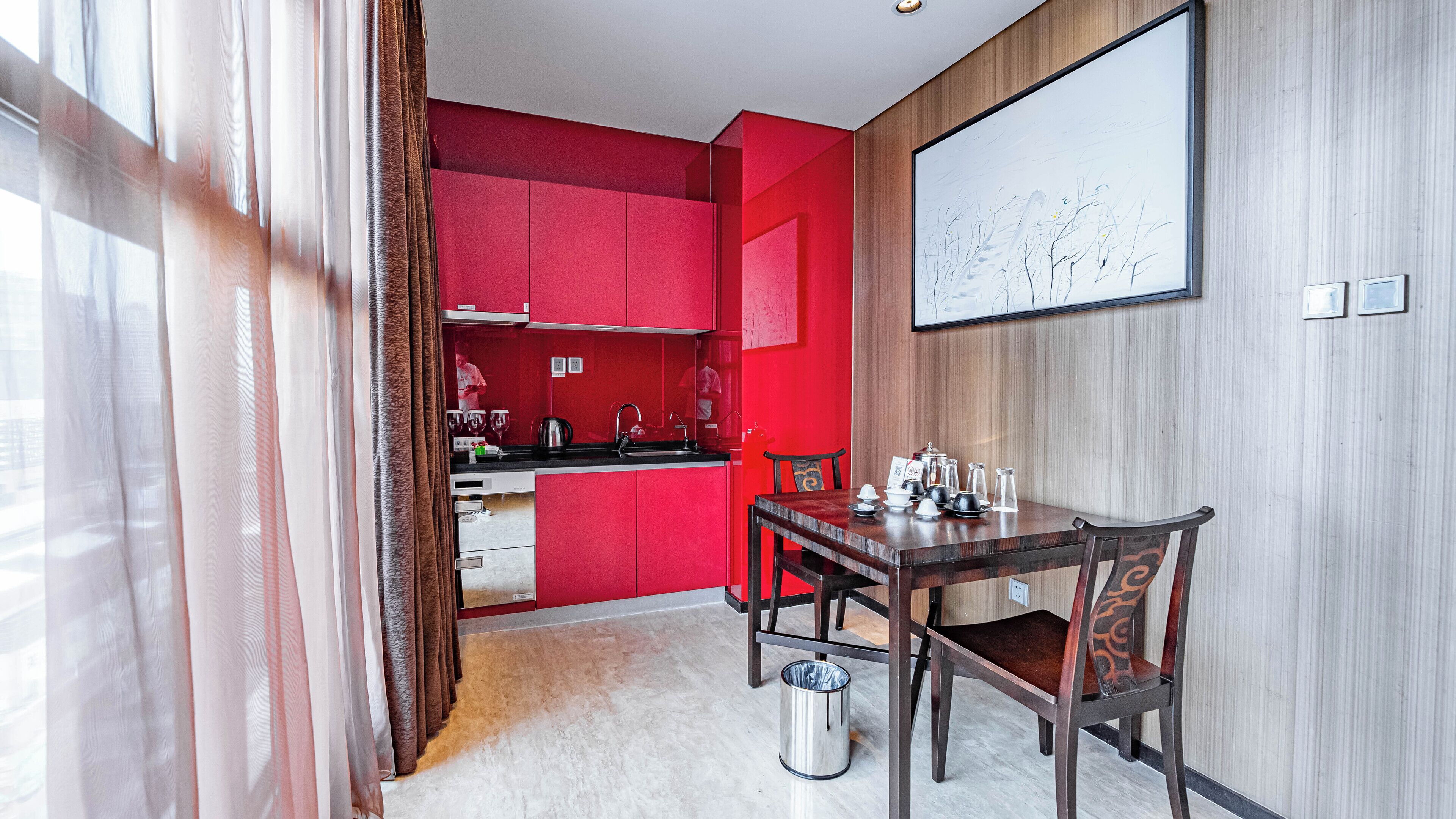 A modern hotel room with a red accent wall, a dining area with a wooden table and chairs, a kitchen area with red cabinets, and a large framed artwork on the wall.