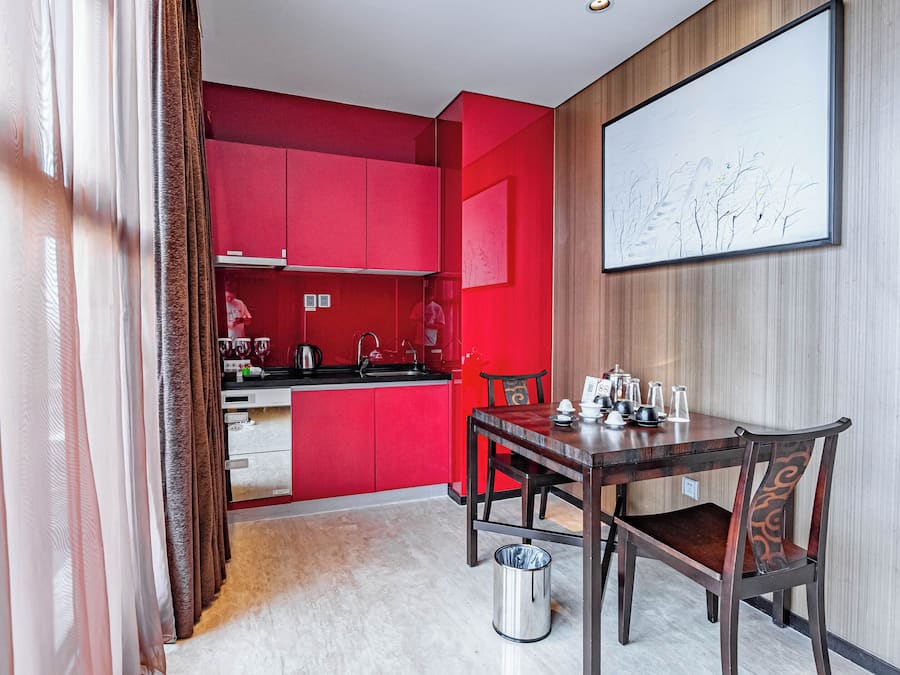 A modern hotel room with a red accent wall, a dining area with a wooden table and chairs, a kitchen area with red cabinets, and a large framed artwork on the wall.