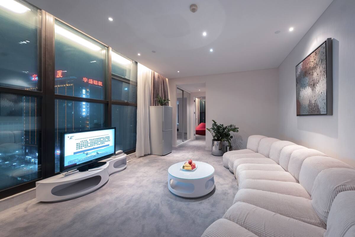 A modern living room with a large flat-screen TV, a round coffee table with fruits, a white sofa, and a view of the city through large windows.