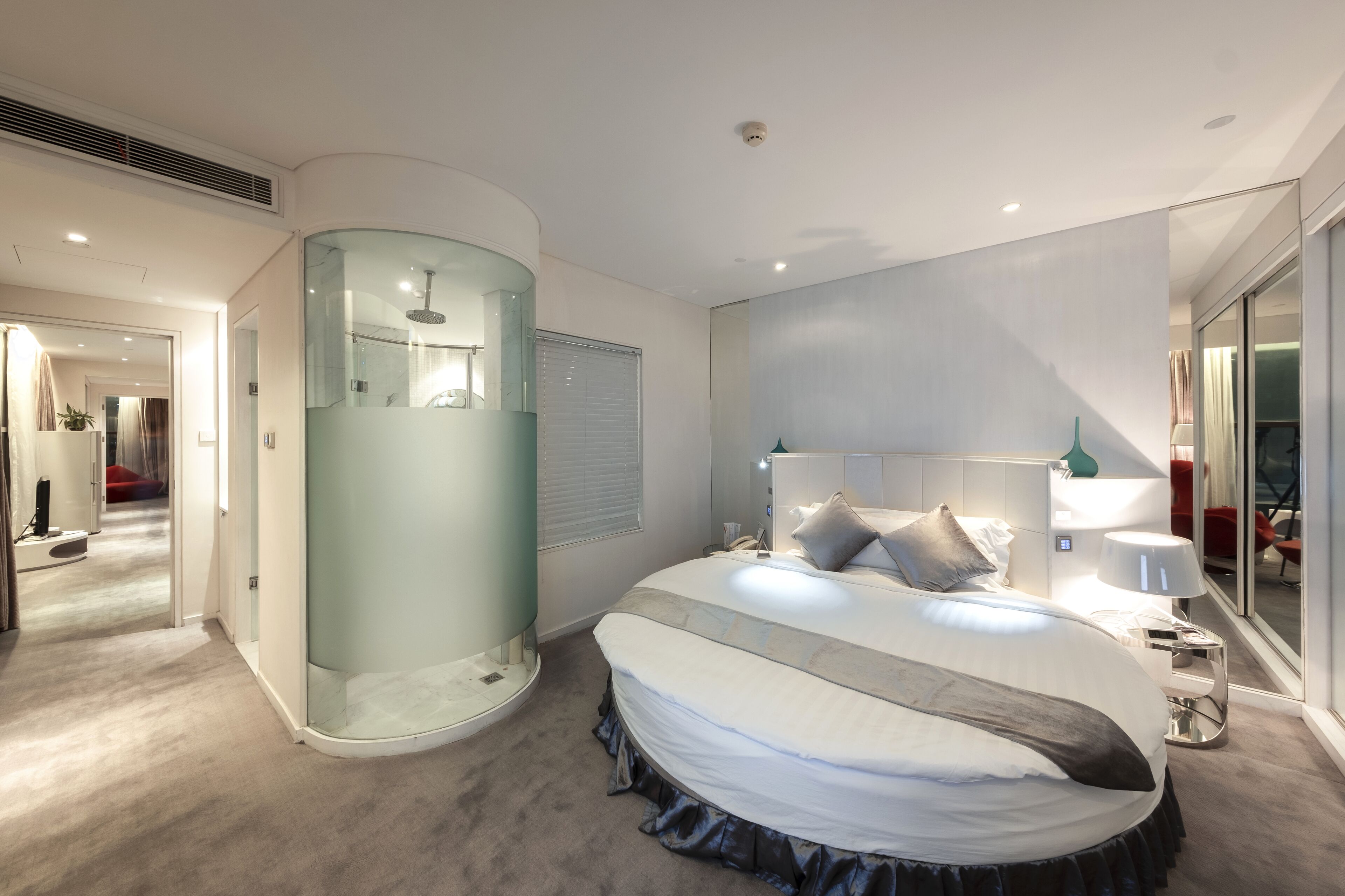 A modern bedroom with a curved glass shower enclosure, a large bed with a skirted base, and a bedside table with a lamp.
