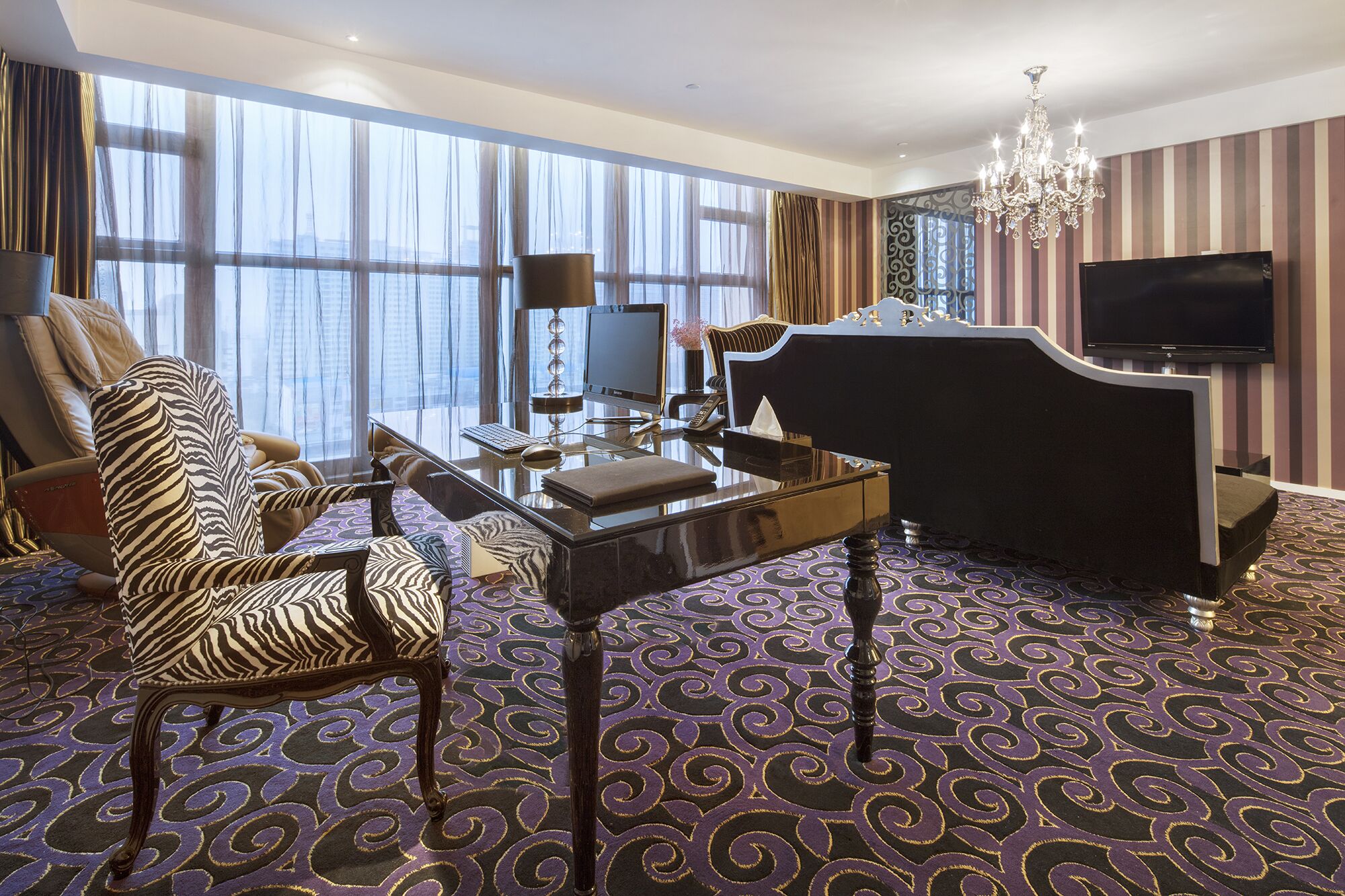 Habitación de hotel con un ventanal grande, un escritorio con computadora, un sofá estampado con motivos de cebra, un cabecero negro y una lámpara de araña.