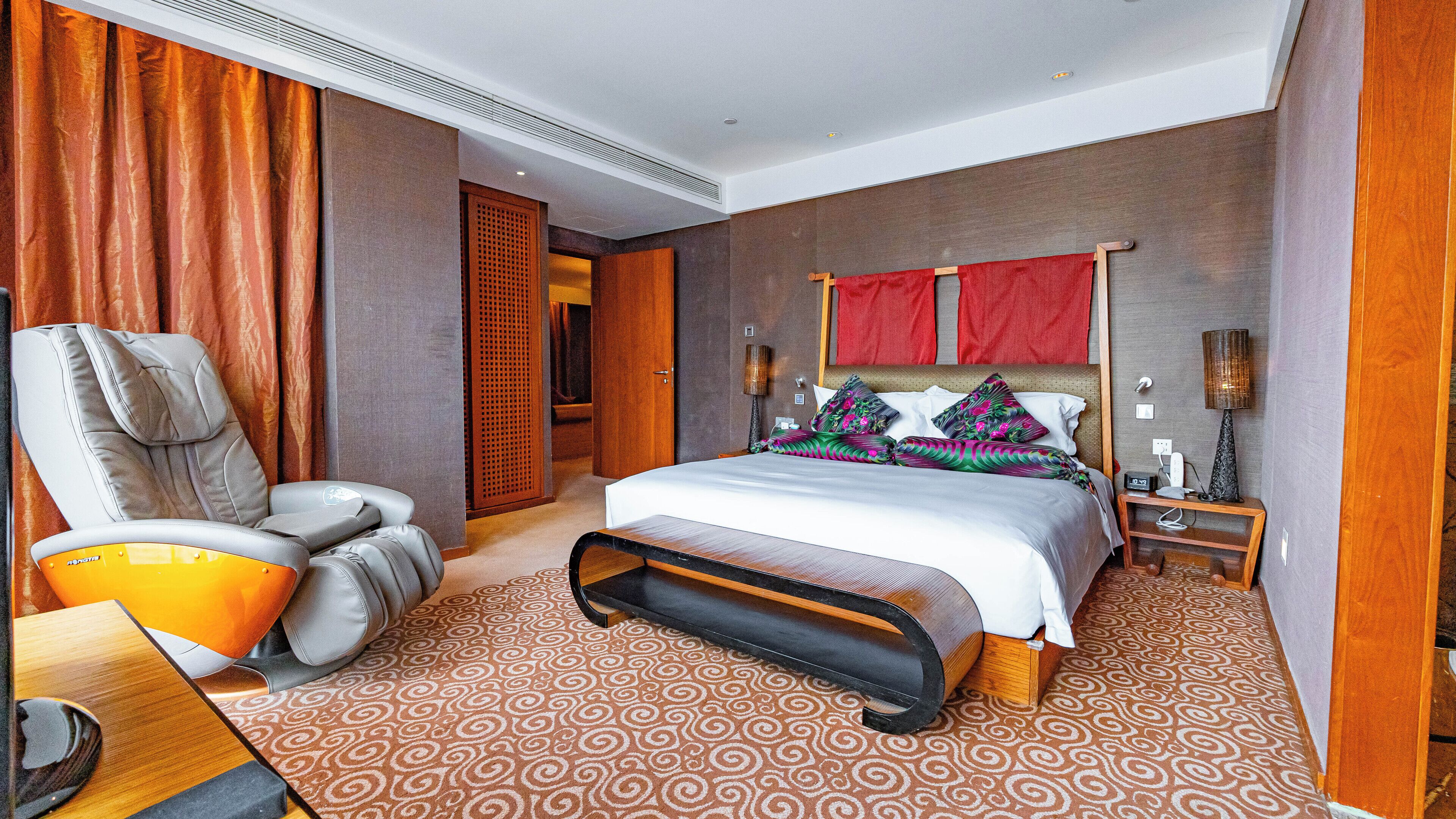 Een hotelkamer met een groot bed, een massagestoel, een nachtkastje met een lamp en uitzicht op de gang.