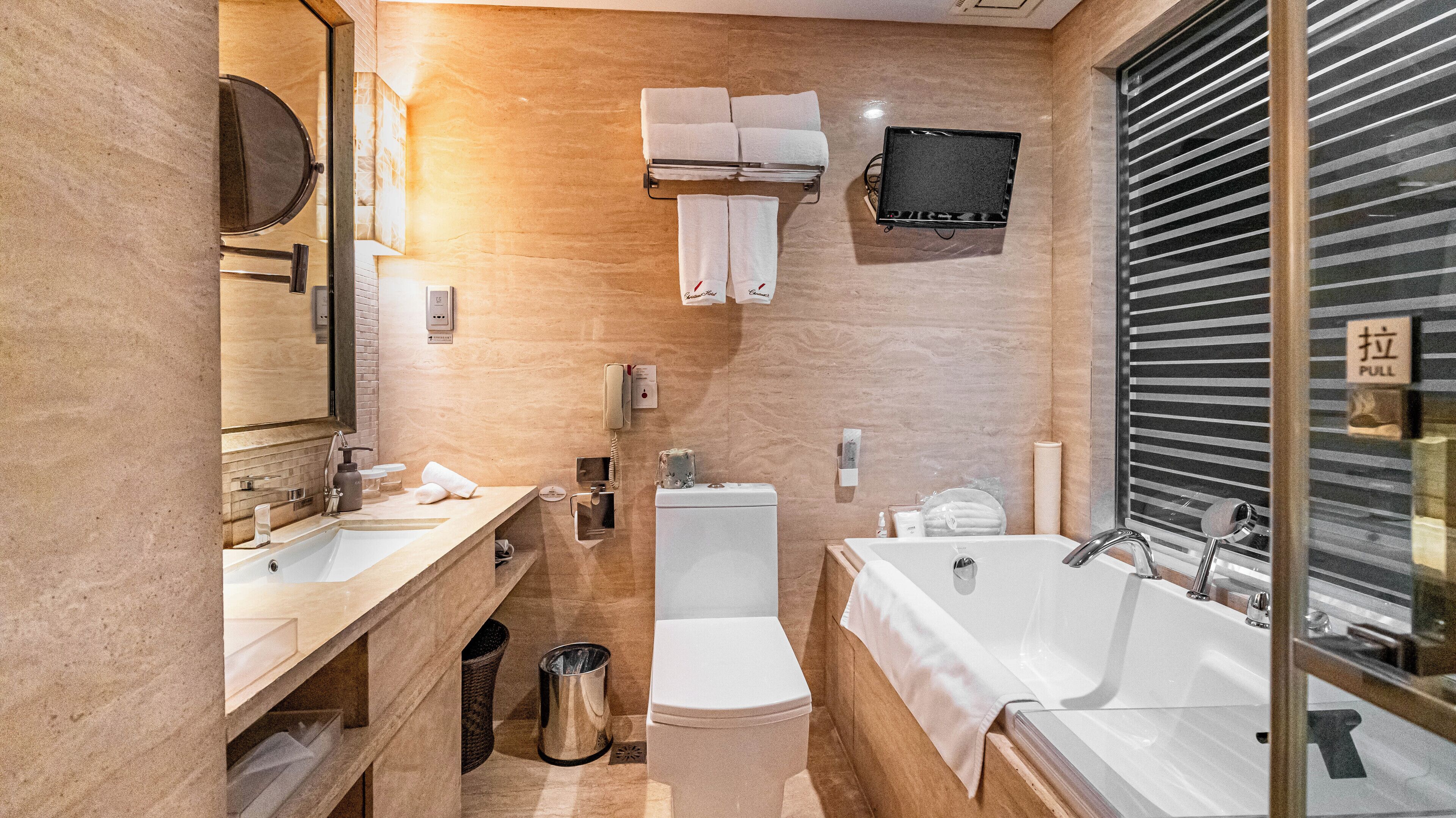 Ein modernes Badezimmer mit einer großzügigen Badewanne, Toilette und Waschbecken. Es gibt einen Spiegel, einen Handtuchhalter und ein Fenster mit Jalousien.