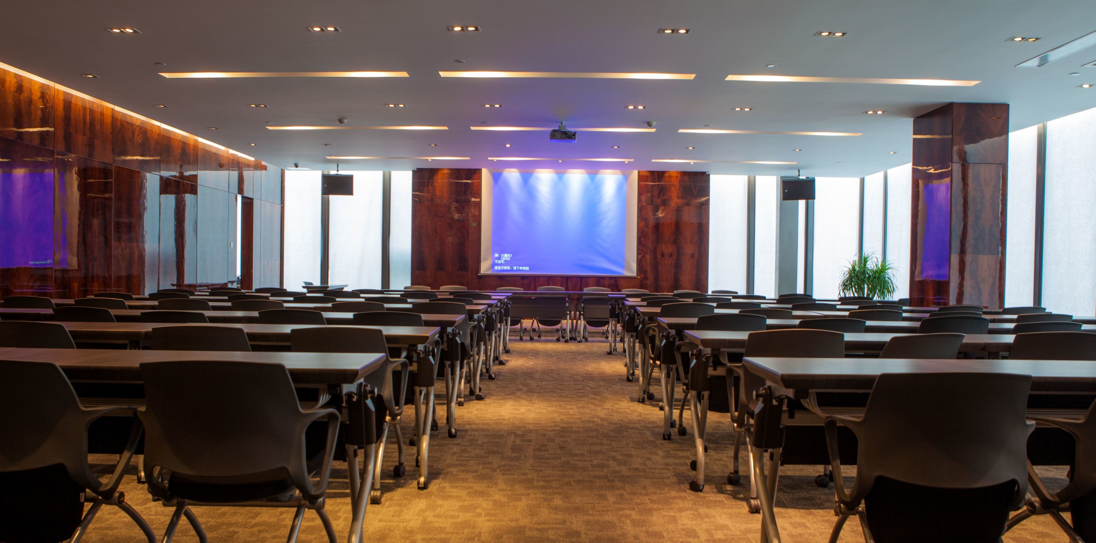 Una sala conferenze con file di tavoli e sedie, un grande schermo di fronte e un moderno design del soffitto.