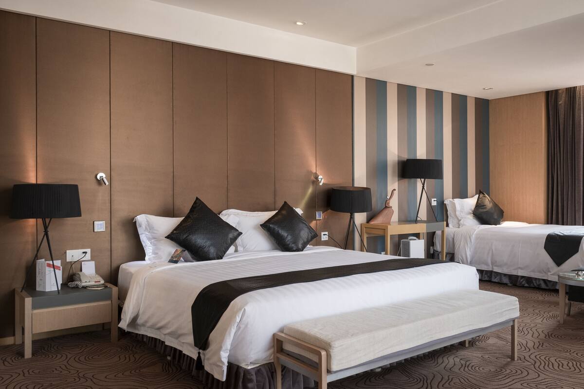 A hotel room with two beds, a bench, bedside tables, lamps, and a wall with vertical stripes.