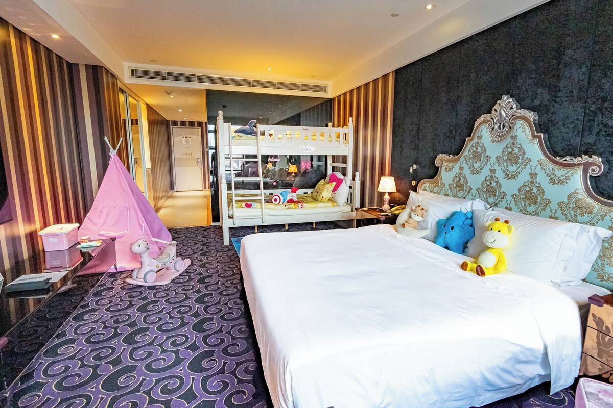 A hotel room with a large bed, a pink play tent, and a bunk bed in the background.