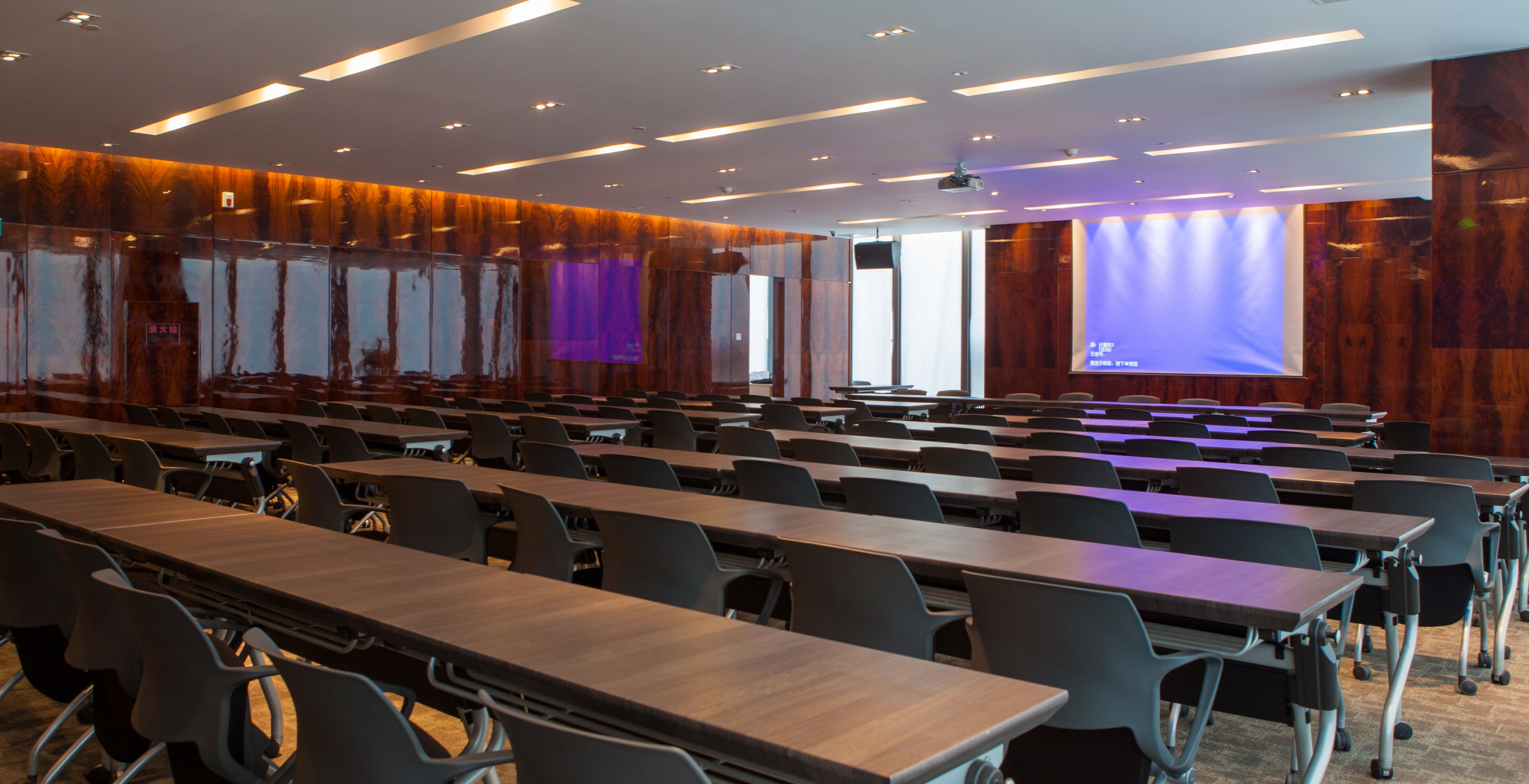 Un salón de conferencias con filas de mesas y sillas, ventanas amplias y una pantalla de presentación.