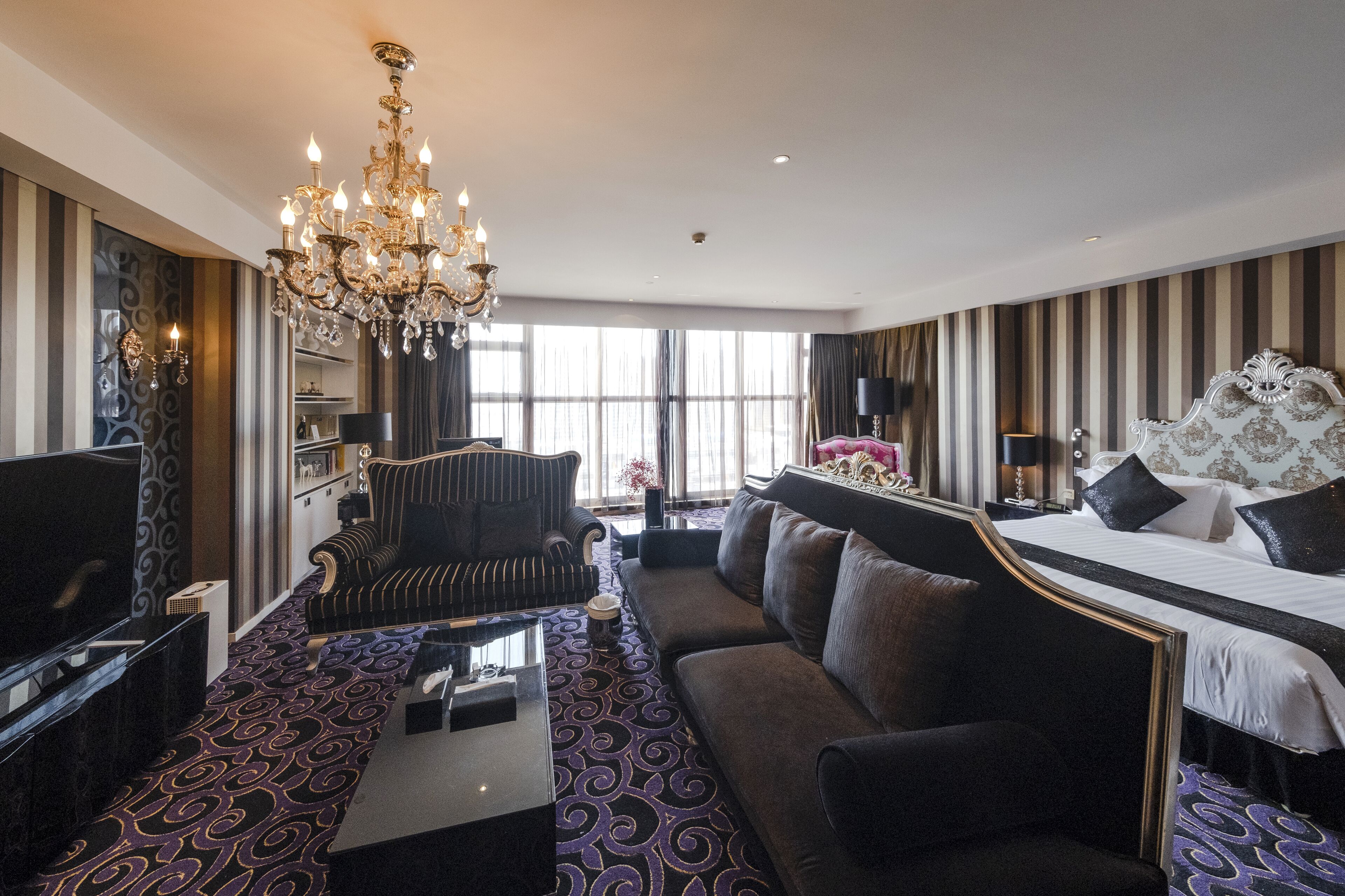 A hotel room with a large bed, a sofa, a TV, a chandelier, and a patterned carpet.