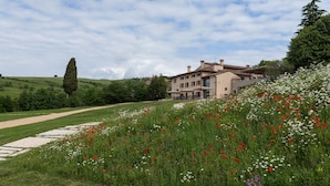 Un edificio grande de dos pisos con un estilo arquitectónico tradicional, rodeado por un paisaje exuberante con diversas flores silvestres y un cielo despejado.