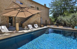 Une piscine entourée de bâtiments en pierre et d’arbres, avec des chaises longues et un grand parasol.