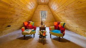A cozy attic room with wooden walls, two colorful armchairs, a small table with a bottle and a lamp, and a window at the far end. - Casa Moritz (Brasov)