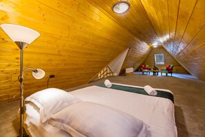 Une pièce triangulaire en bois, chaleureuse et accueillante, avec un lit, une lampe et un petit coin salon agrémenté de chaises colorées.