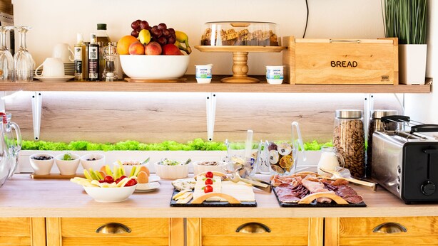 A kitchen counter with a variety of food items, including fruits, cheeses, and meats, and a bread box labeled "BREAD". - Casa Moritz (Brasov)