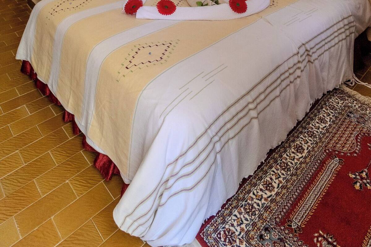 A bedroom with a bed, a ceiling fan, a wall-mounted air conditioner, a patterned rug, and a decorative wall hanging.