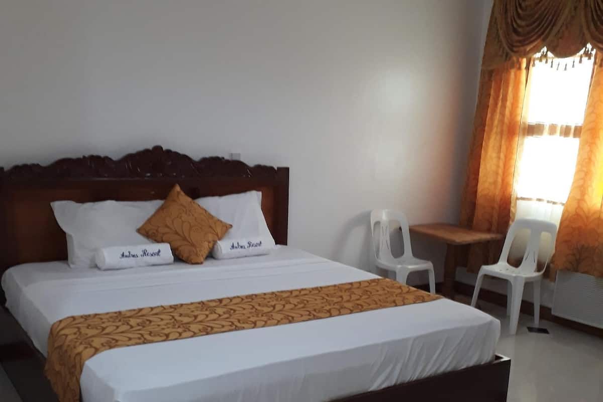 A neatly made bed with a wooden headboard, white bedding, and a patterned throw. There is a small table and two chairs in the corner, and a window with curtains.