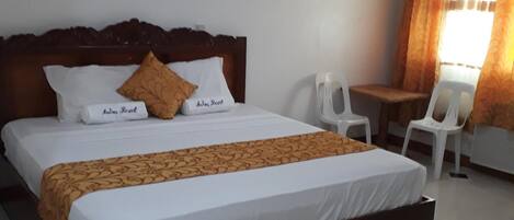 Una cama bien hecha con cabecera de madera, ropa de cama blanca y una manta estampada. En la esquina hay una mesita y dos sillas, y una ventana con cortinas.