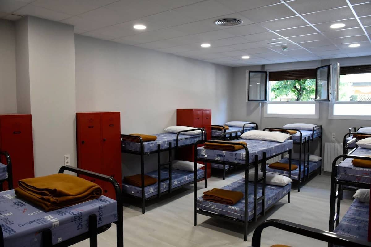 A dormitory room with bunk beds, red lockers, and a window with a view of greenery.