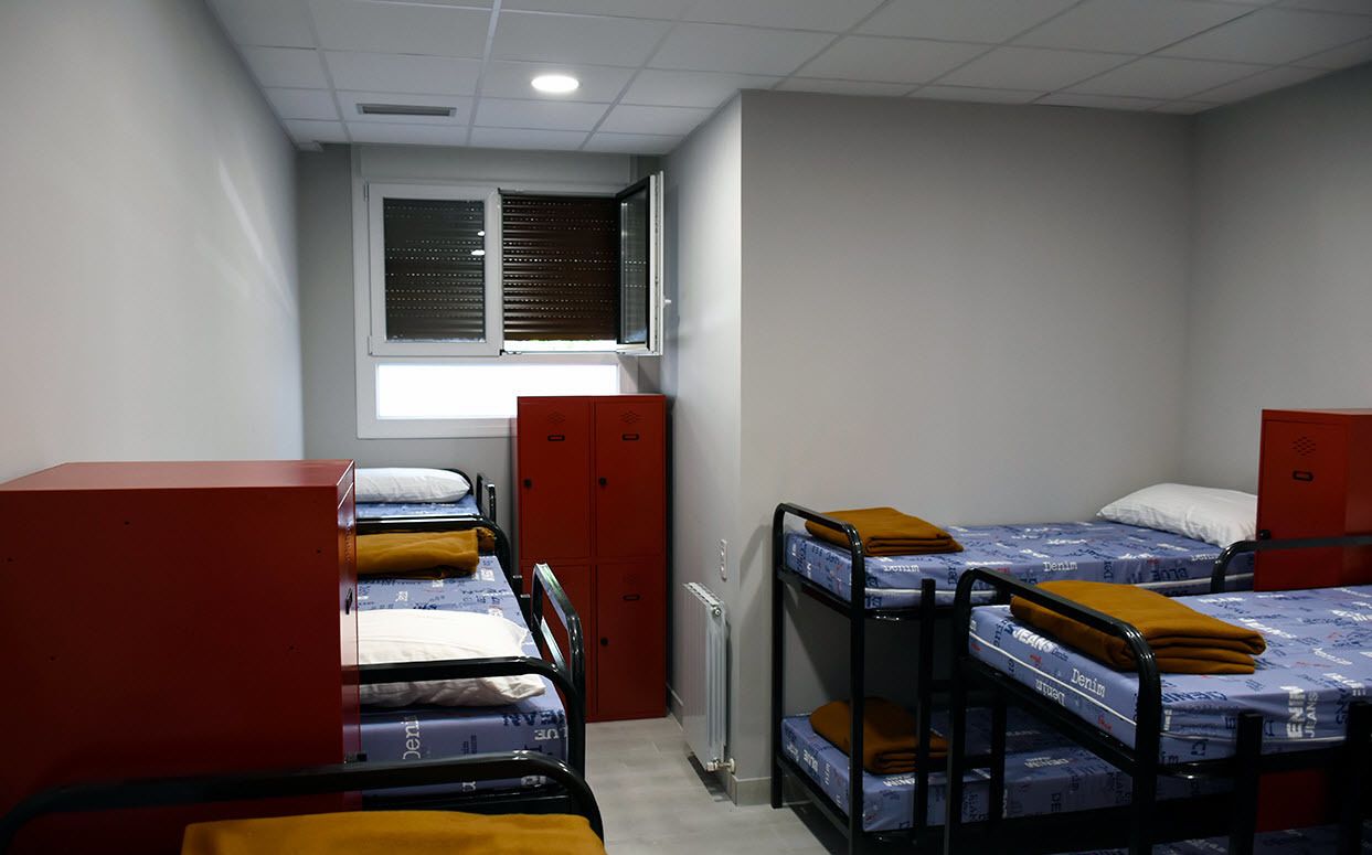 A dormitory room with bunk beds, a radiator, a window with blinds, and a red storage cabinet.