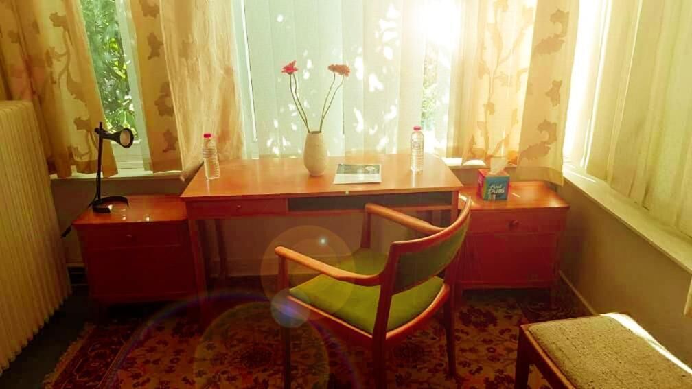 A room with a wooden desk, a chair, a radiator, a window with curtains, and a small table with a bottle and a box.