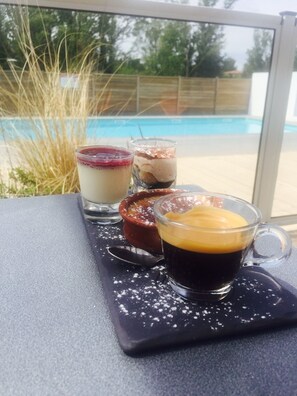A dessert tray with various items including a glass of coffee, a dessert with a creamy topping, a layered dessert, and a dessert with a caramelized top, placed on a table near a window overlooking a pool. - Les Terrasses DAix (Eguilles)