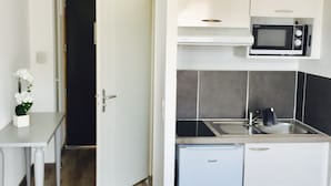 Una cocina compacta con microondas, pileta y heladera pequeña. En primer plano, se ve una mesa blanca con un jarrón de flores.