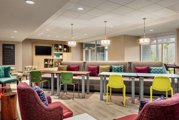 A modern lounge area with a sofa, multiple chairs, a dining table, and pendant lights. - Home2 Suites by Hilton Statesboro (Statesboro)