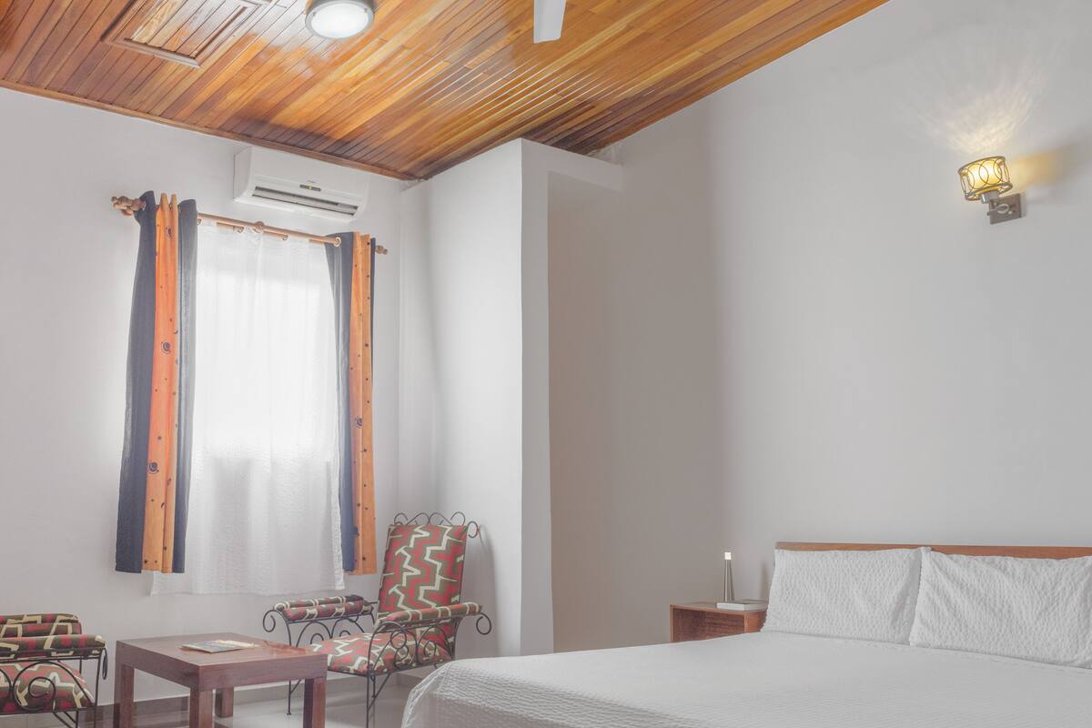 Une chambre avec un plafond en bois, un lit, une table de chevet, une chaise, une table et une fenêtre avec des rideaux.